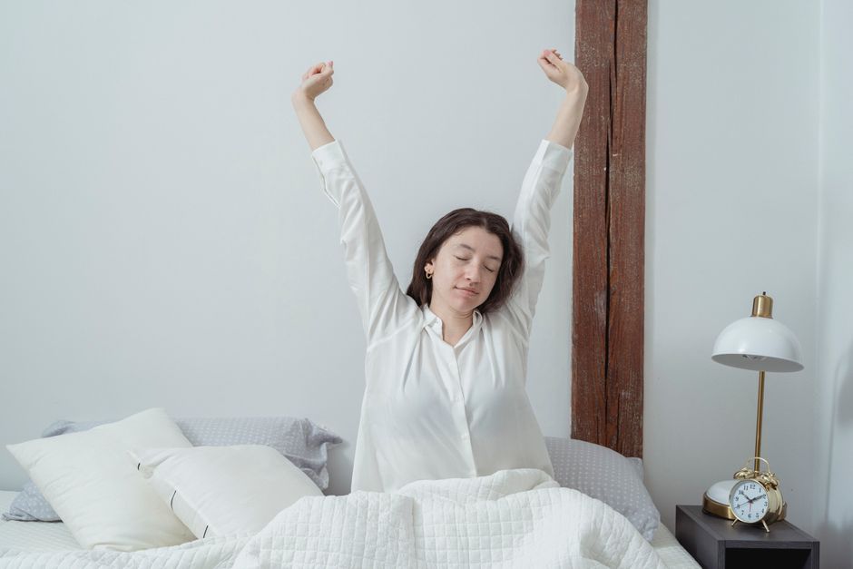 Young brunette with closed eyes sitting on comfortable bed under white blanket and stretching arms up while waking up in morning at home